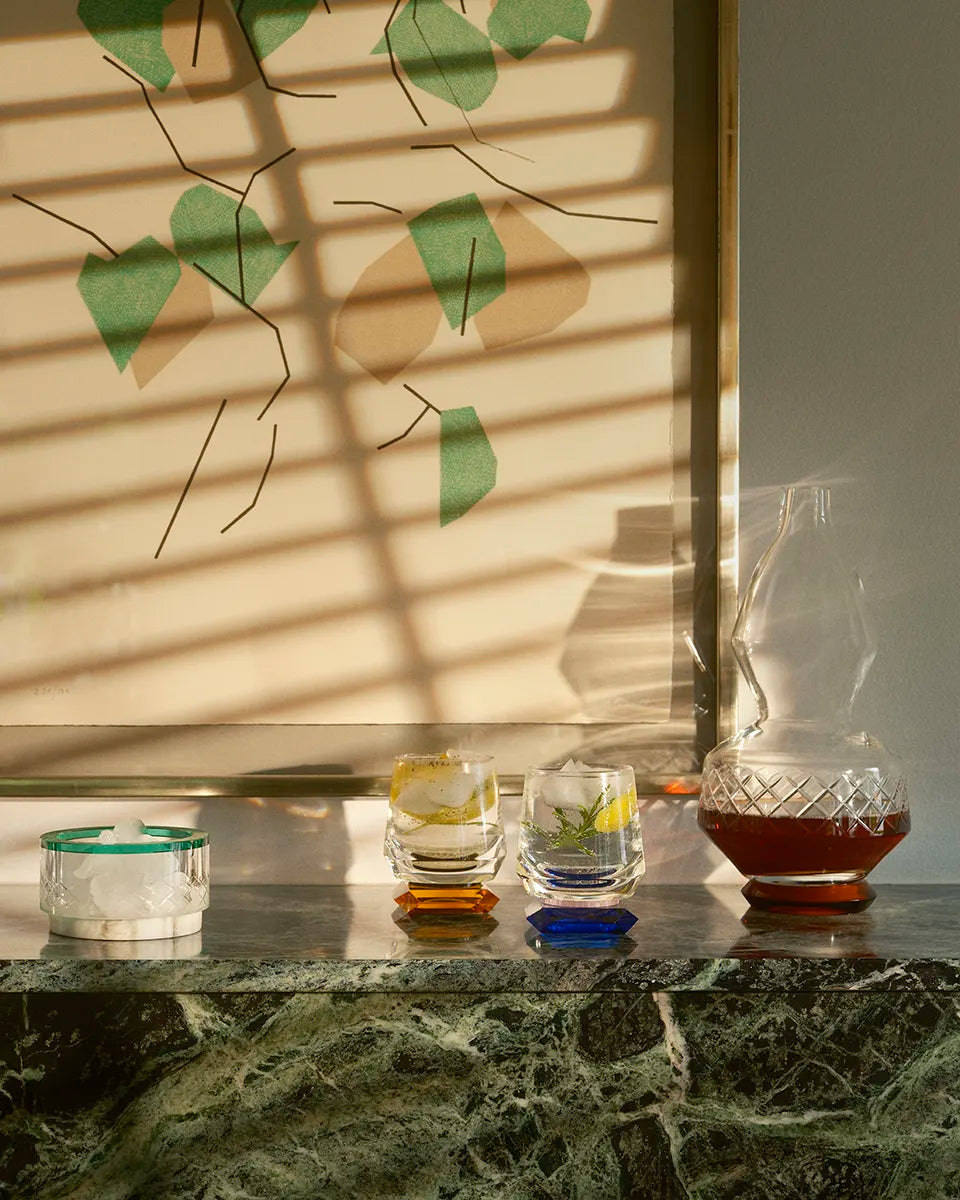 The Whimsy Blue Whisky Glasses rest on a marble surface with a matching crystal ice bowl, casting intricate shadows in golden sunlight.