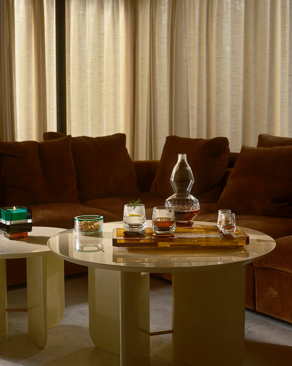 The Soirée Home Scent candle with an emerald green and amber crystal design sits on a round table, surrounded by a whisky set in a cozy living space.