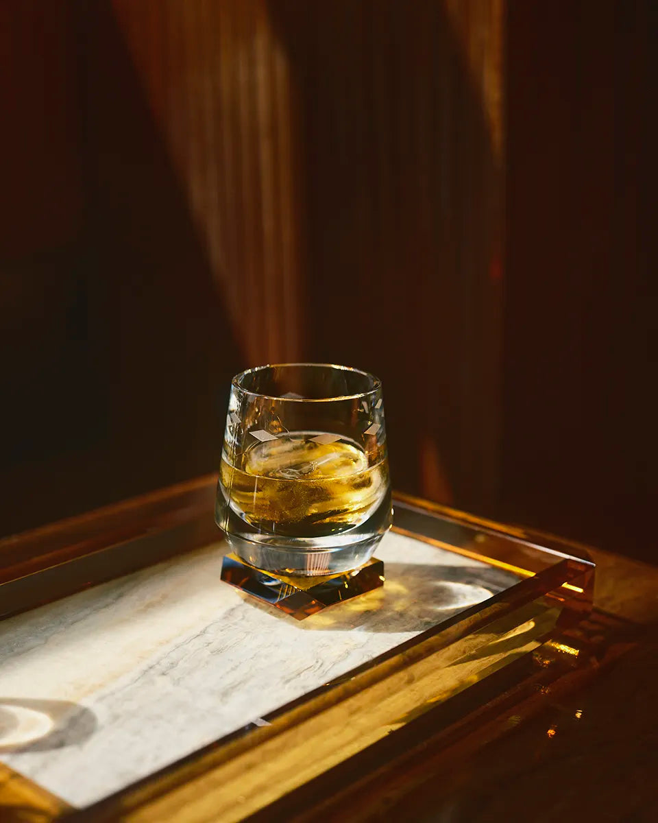 A Whimsy Amber Whisky Glass filled with golden whisky sits on a reflective amber tray, bathed in moody, warm lighting.